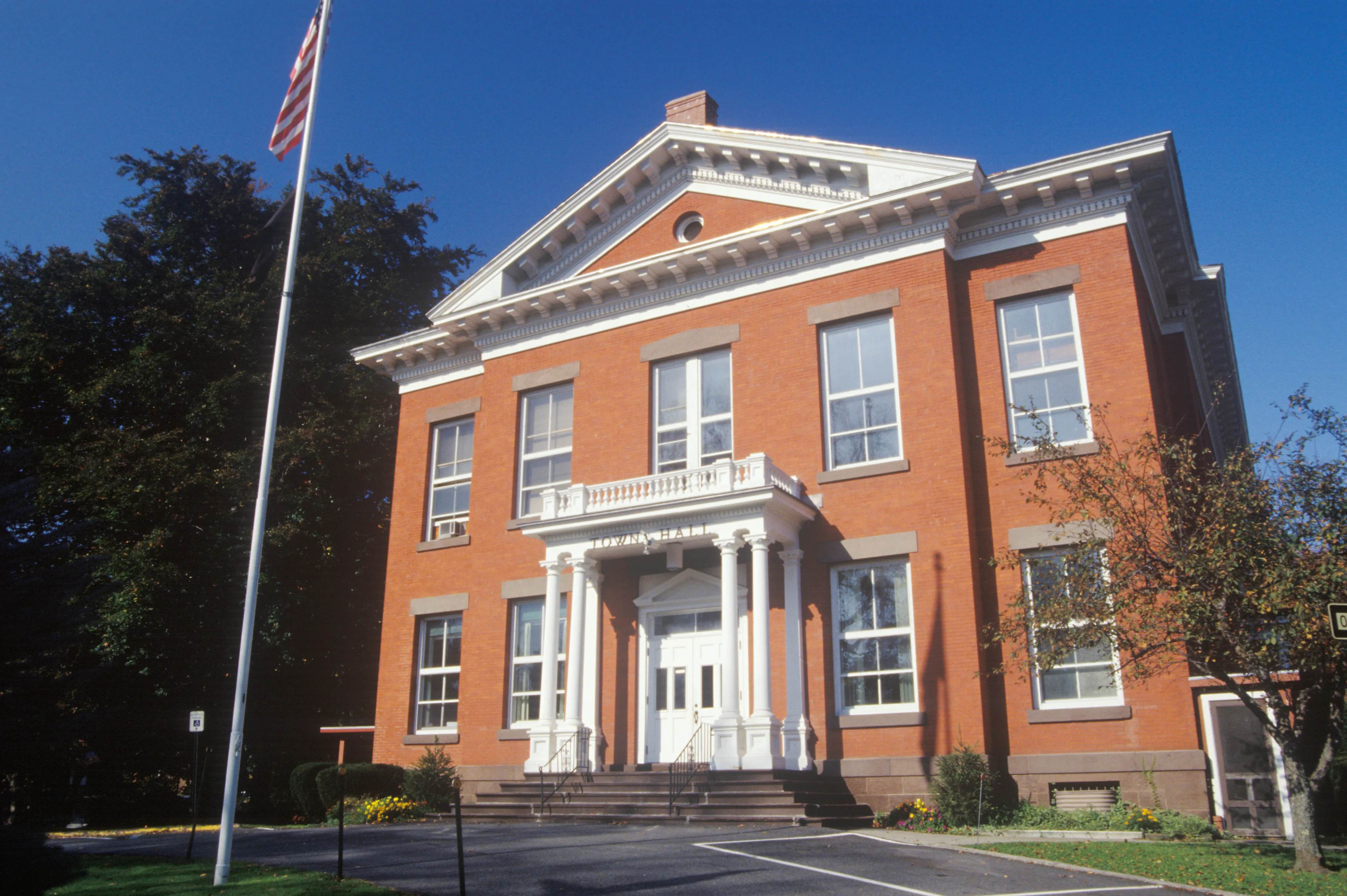 Historic brick courthouse building with white columns, pediment, and front entrance portico. American flag flies on left side under clear blue sky with autumn foliage.