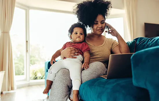 Mother holding toddler while working on laptop on blue couch at home with large windows in background