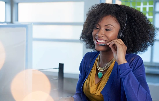 Professional woman wearing blue blazer and yellow scarf smiling while talking on phone at modern office desk with computer monitor