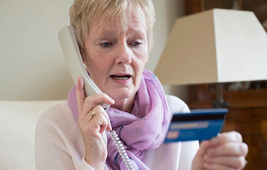 Elderly woman with short white hair wearing a purple cardigan speaking on a corded telephone while holding a blue credit card